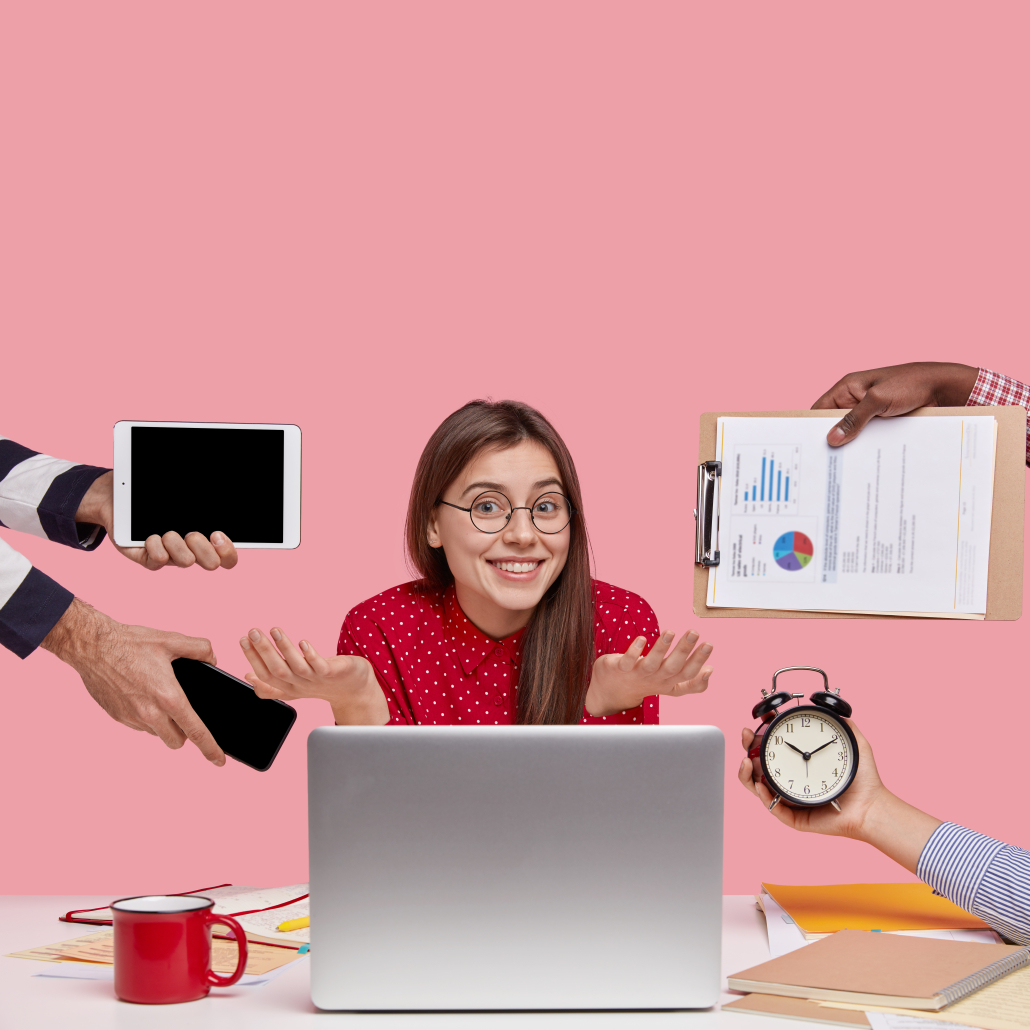 Smiling beautiful dark haired young woman spreads palms, smiles broadly, has uncertain facial expression, uses laptop computer, prepares project work, surrounded with modern gadgets and papers